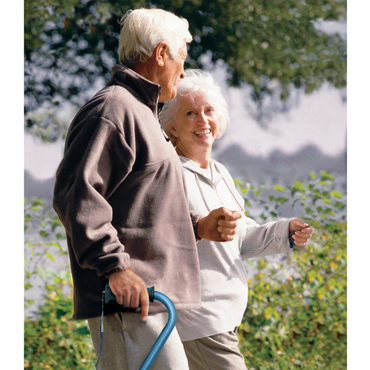 An older couple strolls hand in hand, smiling amid green trees. The man uses a Drive Airgo Comfort-Plus Adjustable Quad Cane for balance, and both wear warm, casual clothing while enjoying the outdoors together.