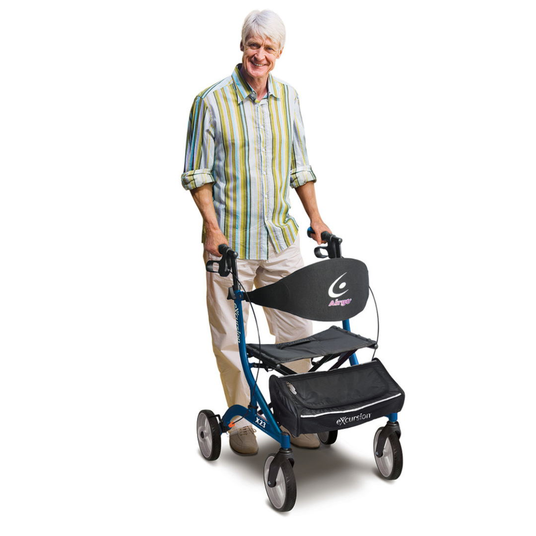 A smiling older adult with gray hair, in a striped shirt and beige pants, stands behind a Drive Airgo eXcursion X23 Lightweight Rollator for Tall Users, featuring a black seat and storage pouch, against a white background.