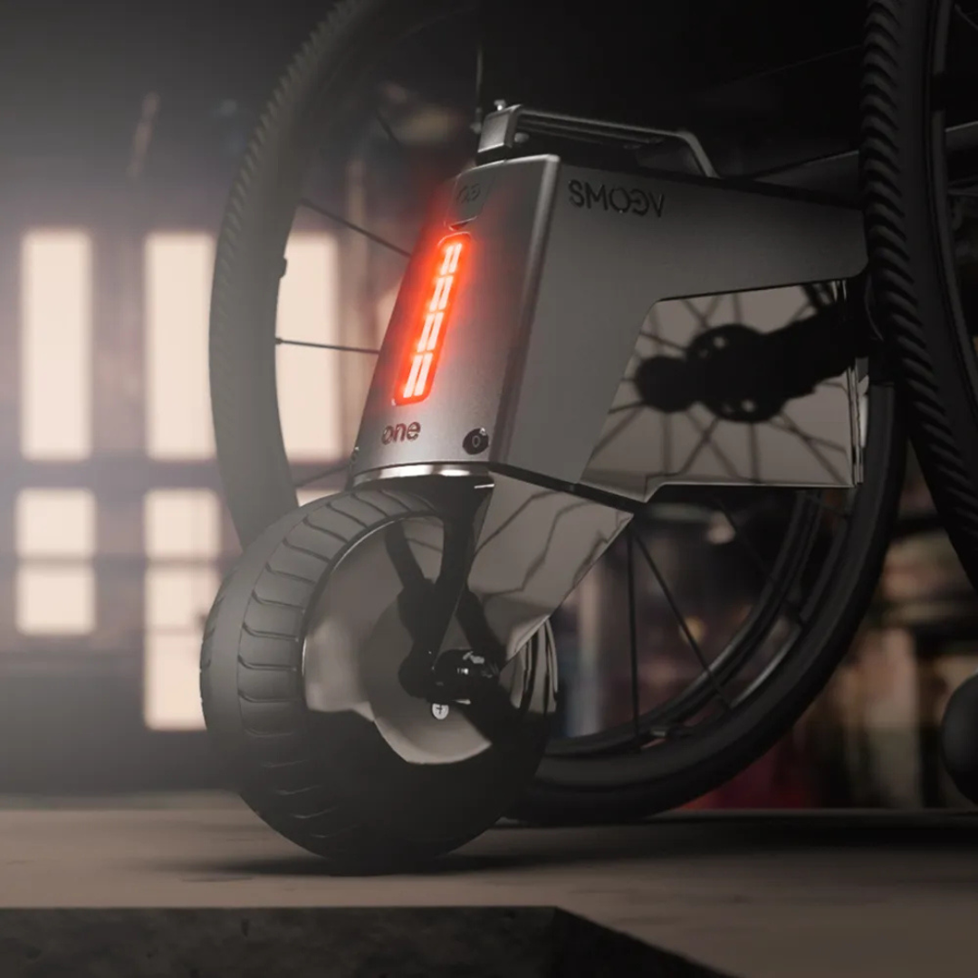 Close-up of the Alber SMOOV One Power Assist, a modern motorized wheelchair attachment, displaying speed 2.0 on its digital screen while connected to the front wheel against a softly lit indoor background.
