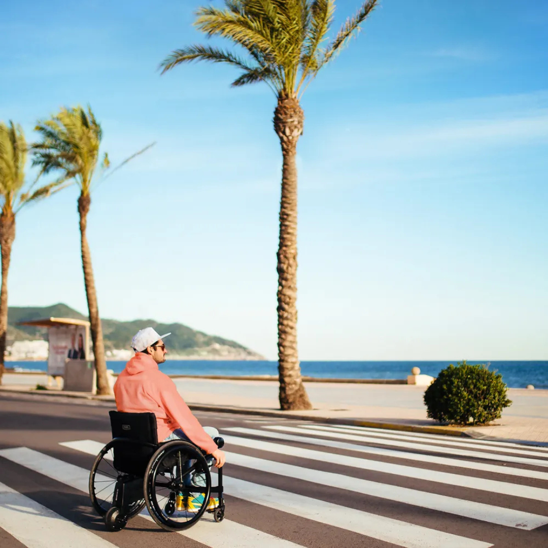 A man in a wheelchair with the Alber SMOOV One Power Assist, wearing a pink hoodie and white cap, crosses a striped crosswalk near the beach, with tall palm trees and blue sky in the background.