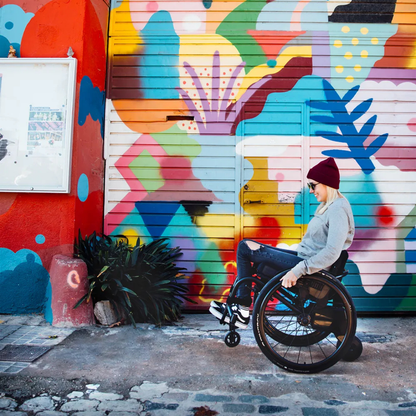 A person confidently does a wheelie in a wheelchair with the Alber SMOOV One Power Assist on a cobblestone sidewalk in front of a colorful mural, wearing a gray sweater, jeans, and red beanie.