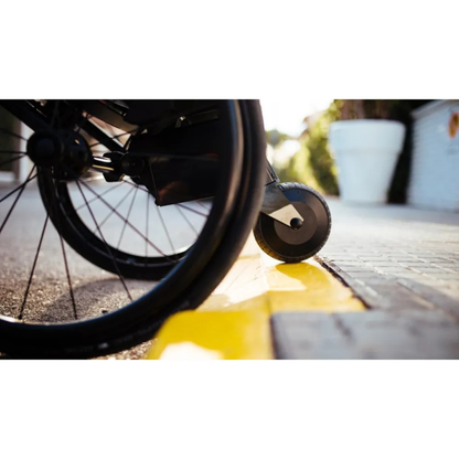 A close-up shows a wheelchair wheel fitted with the Alber SMOOV One Power Assist on a sidewalk nearing a yellow curb in bright sunlight, highlighting accessibility challenges faced by wheelchair users.