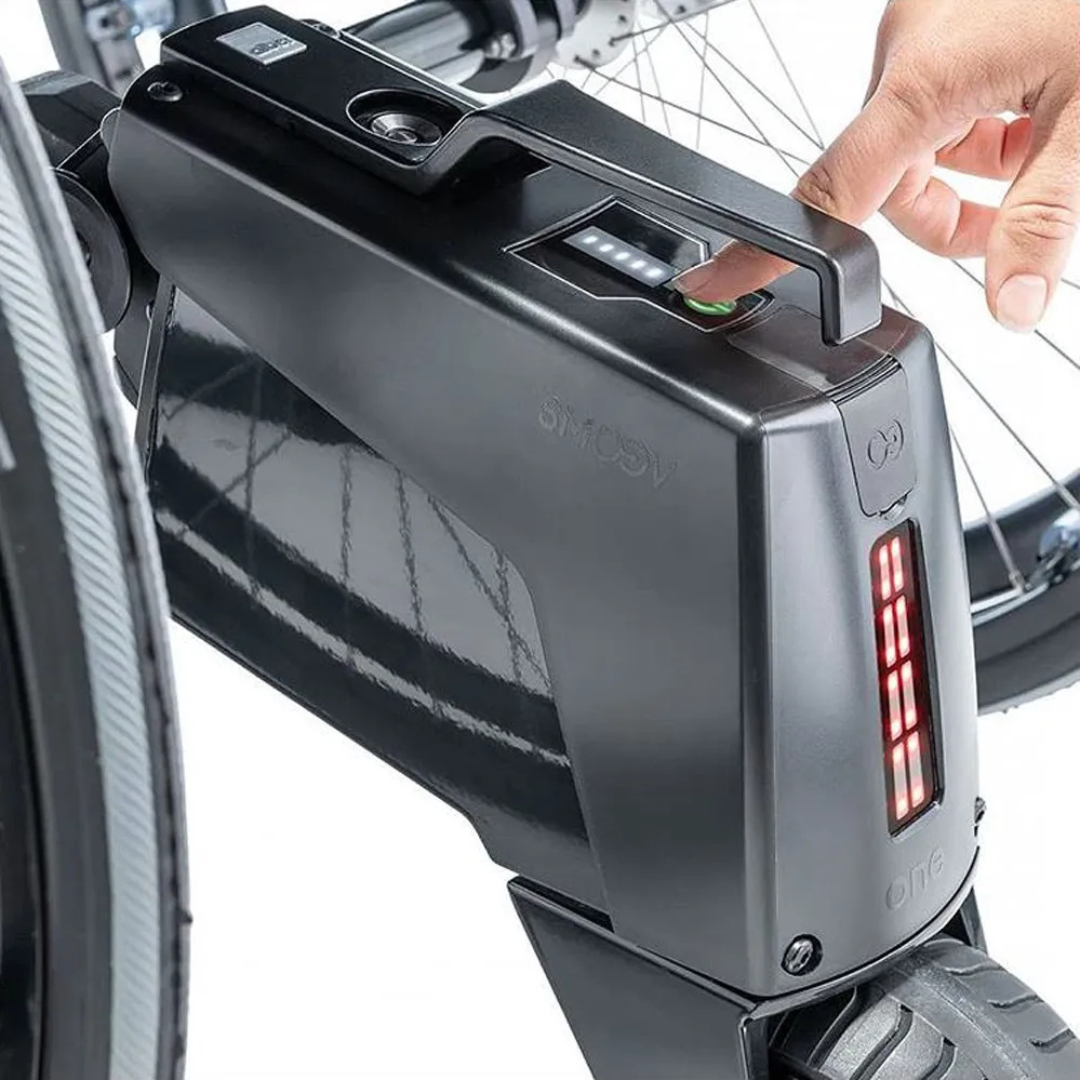 A hand presses a fingerprint scanner on a black Alber SMOOV One Power Assist e-bike battery attached to a bike frame. Red LEDs glow as the battery, equipped with a sturdy handle, enables easy removal and electric drive power assist.