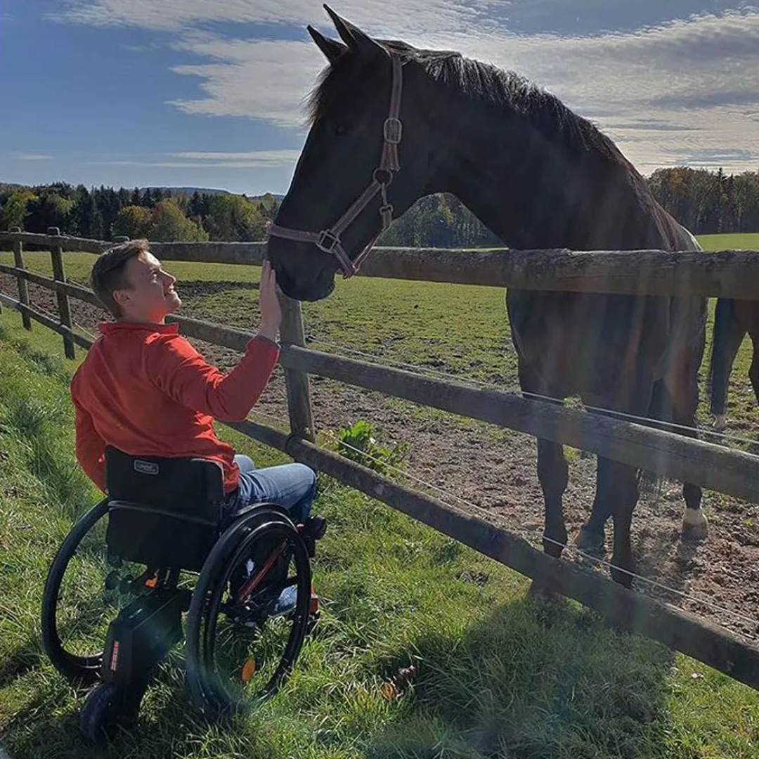 A person in a wheelchair equipped with the Alber SMOOV One Power Assist gently reaches out to touch a black horse over a wooden fence in a sunny, grassy field with trees in the background.