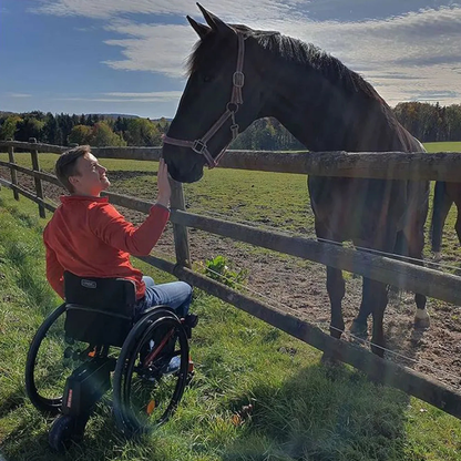A person in a wheelchair equipped with the Alber SMOOV One Power Assist gently reaches out to touch a black horse over a wooden fence in a sunny, grassy field with trees in the background.