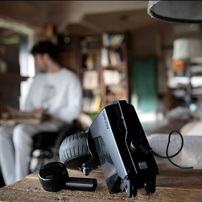 A close-up of the Alber SMOOV One Power Assist joystick and a wireless earbud on a table, while a blurred person in white sits in a cozy, well-lit room with bookshelves.