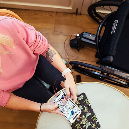A person in a pink sweatshirt sits at a round table using a smartphone with a floral case. Part of an Alber SMOOV One Power Assist wheelchair is visible beside them on the wooden floor.