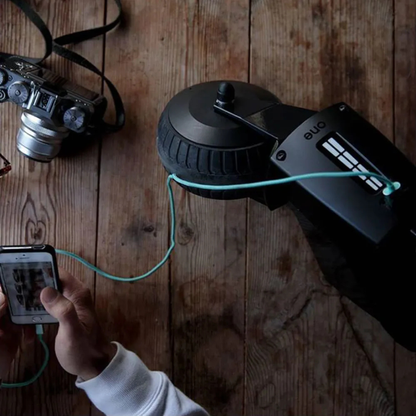 A person charges their smartphone with the Alber SMOOV One Power Assist, hooked up to a tire-shaped device, while a camera rests on a rustic wooden table nearby.