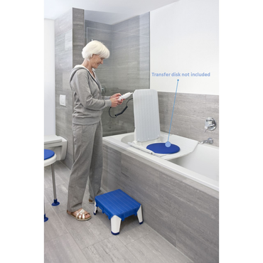 An older woman in a grey tracksuit stands by a bathtub with an Aquatec XL Heavy Duty Reclining Bath Lift inside. A blue transfer disk labeled “not included” is on the lift seat, and a blue step stool is in front of the tub.