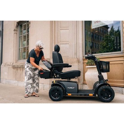 An older woman with short gray hair, in a black shirt and patterned pants, stands on a sidewalk placing essentials into the key-locking Pride Mobility Scooter Storage Pod attached to her black mobility scooter.