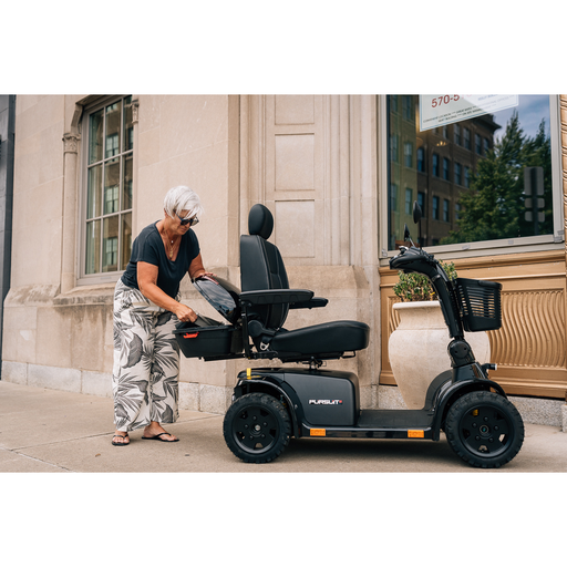 An older woman with short gray hair, in a black shirt and patterned pants, stands on a sidewalk placing essentials into the key-locking Pride Mobility Scooter Storage Pod attached to her black mobility scooter.