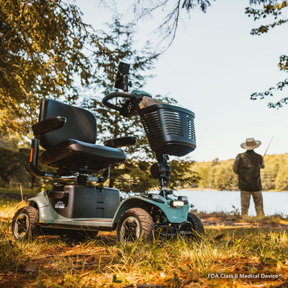 The Pride Baja® Bandit 4 Wheel Scooter, known for its rugged off-road capabilities and full suspension, is parked on grass near a lake. A person in a wide-brimmed hat fishes nearby, surrounded by trees and water, creating a serene outdoor setting.