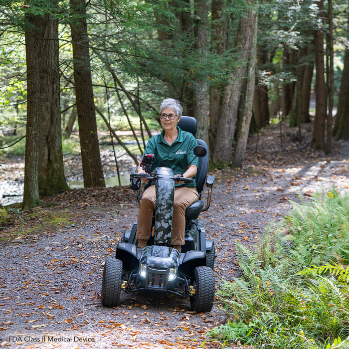 A person with short gray hair, wearing glasses and a green shirt, rides a Pride Baja® Wrangler® 2 on a forest path surrounded by trees and ferns. The scooters CTS Suspension smoothly navigates the fallen leaves on the trail, highlighting this rugged autumn terrain.