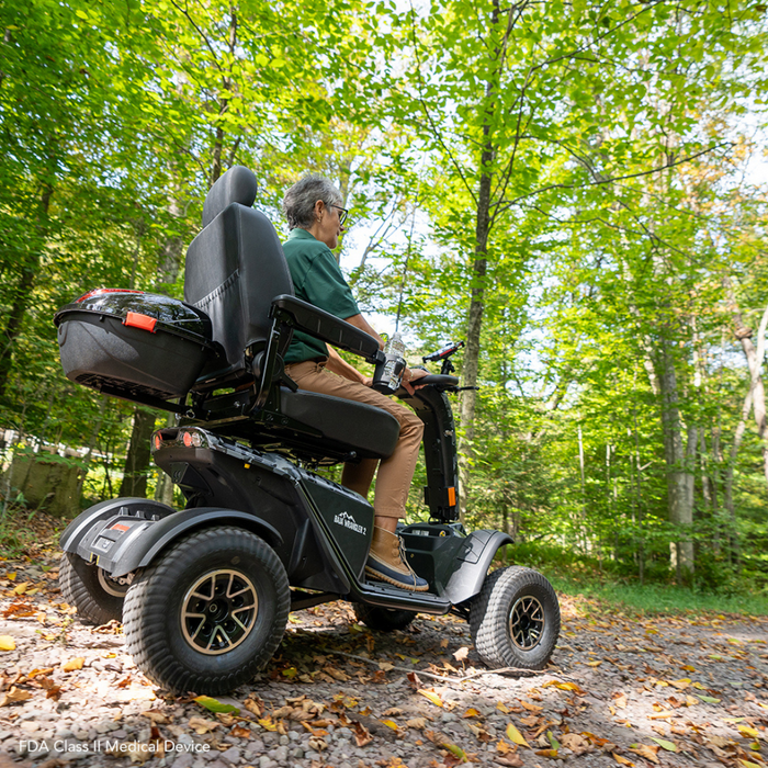 A person navigates a forest trail on the robust Pride Baja® Wrangler® 2, an all-terrain outdoor mobility scooter by Baja Mobility. Surrounded by bright green trees and fallen leaves, it features CTS Suspension for adventurous rides.