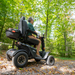 A person navigates a forest trail on the robust Pride Baja® Wrangler® 2, an all-terrain outdoor mobility scooter by Baja Mobility. Surrounded by bright green trees and fallen leaves, it features CTS Suspension for adventurous rides.