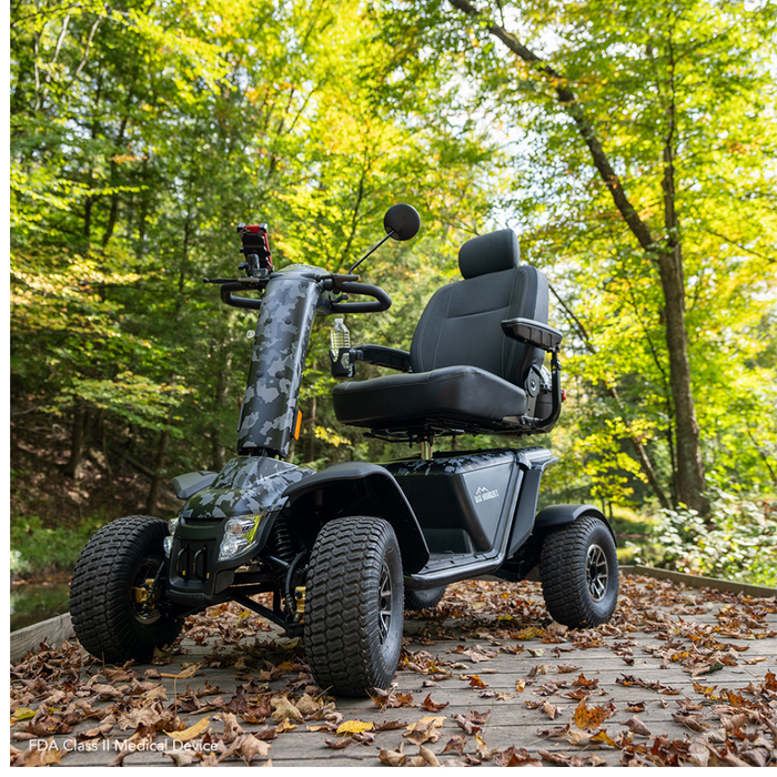 A Pride Baja® Wrangler® 2 Heavy Duty Outdoor Scooter in camouflage is parked on a wooden path in a lush forest. With large tires, armrests, CTS Suspension, and a backrest, it complements the natural backdrop.