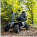 A Pride Baja® Wrangler® 2 Heavy Duty Outdoor Scooter in camouflage is parked on a wooden path in a lush forest. With large tires, armrests, CTS Suspension, and a backrest, it complements the natural backdrop.