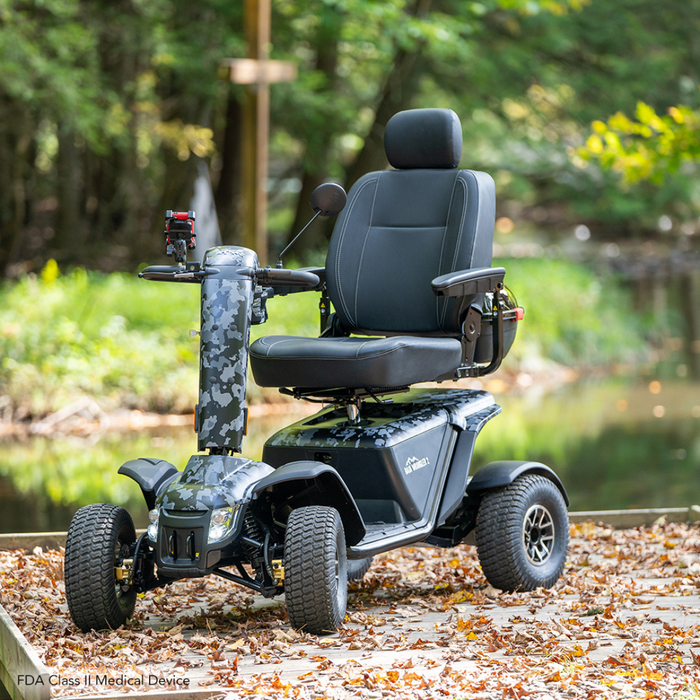 The Pride Baja® Wrangler® 2 Heavy Duty Outdoor Scooter is showcased on a wooden path among fallen leaves, surrounded by lush greenery and a tranquil pond. Built for rugged terrain, it features CTS Suspension, comfortable seating, and sturdy handlebars for a smooth ride through nature.