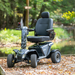 The Pride Baja® Wrangler® 2 Heavy Duty Outdoor Scooter is showcased on a wooden path among fallen leaves, surrounded by lush greenery and a tranquil pond. Built for rugged terrain, it features CTS Suspension, comfortable seating, and sturdy handlebars for a smooth ride through nature.