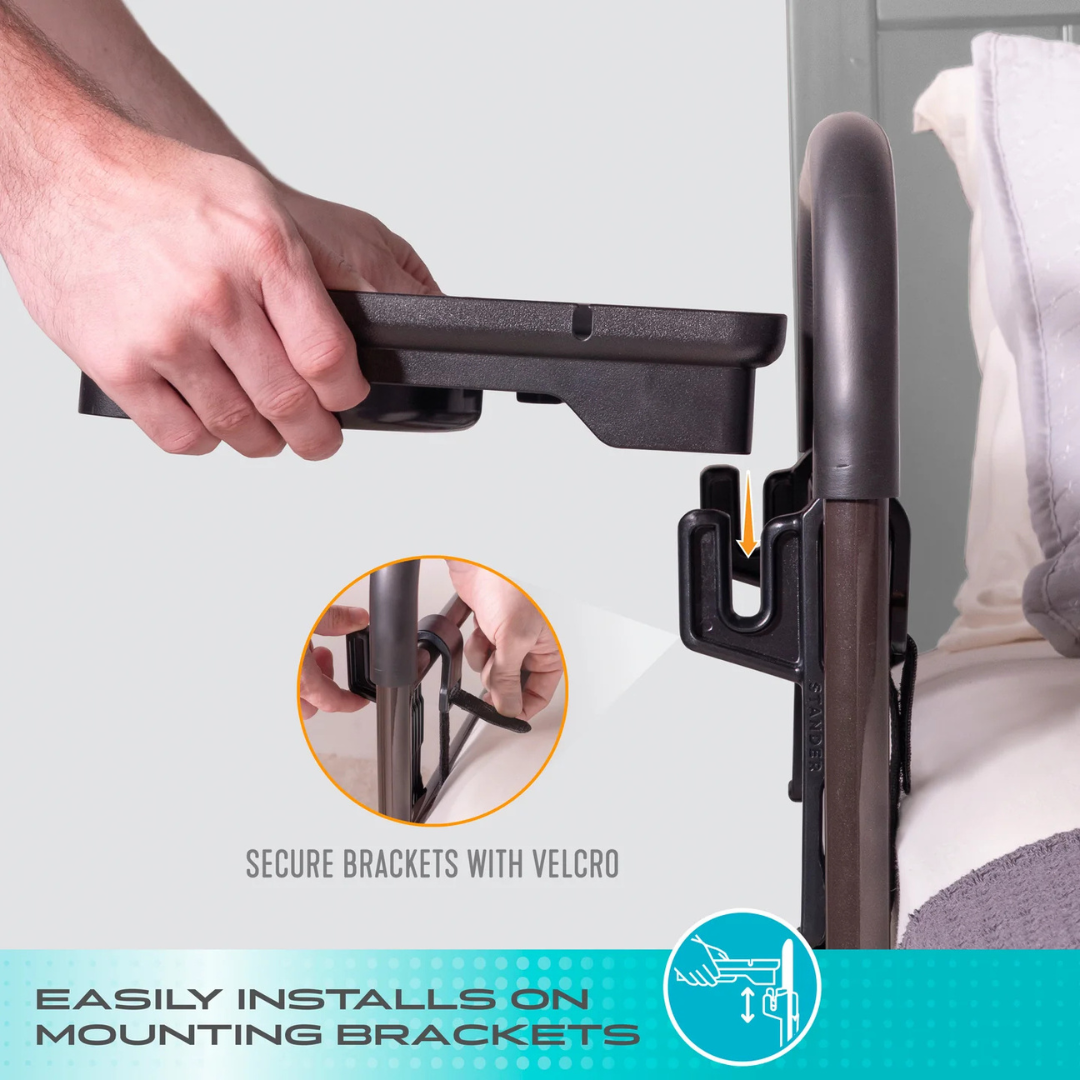 A person’s hand attaches the Stander Bed Rail Tray Accessory (in black) to a bed rail mounting bracket using Velcro, showing simple installation. The text at the bottom reads, Easily installs on Stander Bed Rail mounting brackets.