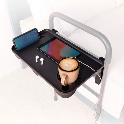 The Stander Bed Rail Tray Accessory, attached to a Stander Bed Rail, secures a smartphone, tablet (with charging cable), wireless earbuds, and a brown coffee mug—keeping essential items conveniently within reach.