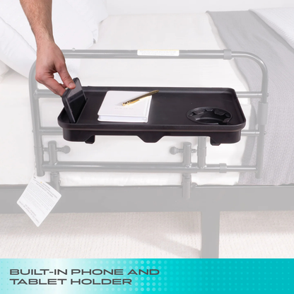 A hand places a phone onto the built-in holder of the black Stander Bed Rail Tray Accessory, attached to a bed rail. The tray holds a notepad, pencil, and cup. Text reads: BUILT-IN PHONE AND TABLET HOLDER—ideal bed rail accessory.