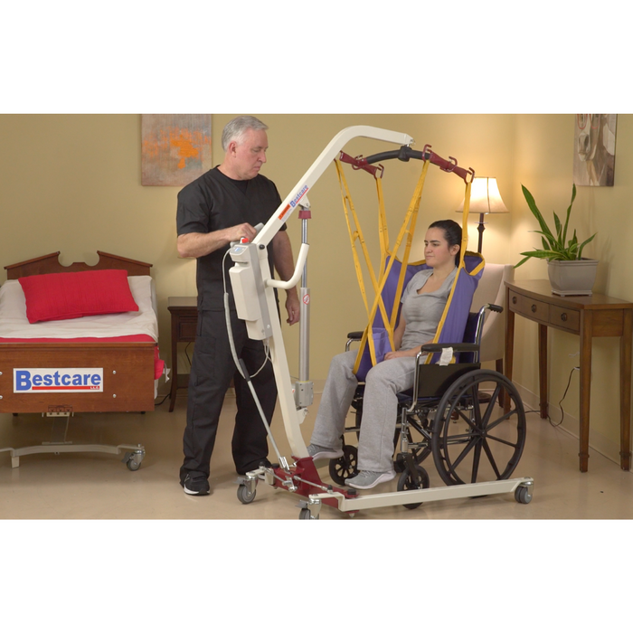 A man uses the Bestcare Bestlift PL182 Full Body Electric Patient Lift to assist a woman in a wheelchair. They are in a room with a hospital bed and side table, designed for bariatric patients comfort.