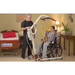 A man uses the Bestcare Bestlift PL182 Full Body Electric Patient Lift to assist a woman in a wheelchair. They are in a room with a hospital bed and side table, designed for bariatric patients comfort.