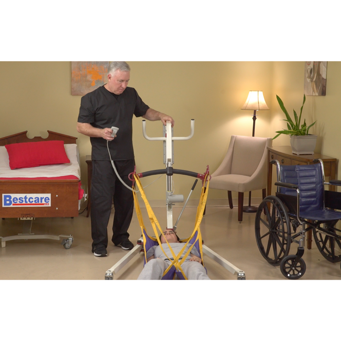 In softly lit surroundings, a caregiver in black scrubs uses the Bestcare Bestlift PL182 Full Body Electric Patient Lift to assist a patient from a sling on the floor. The room features a bed, wheelchair, chair, cozy lamp, and plant adding warmth to the scene.