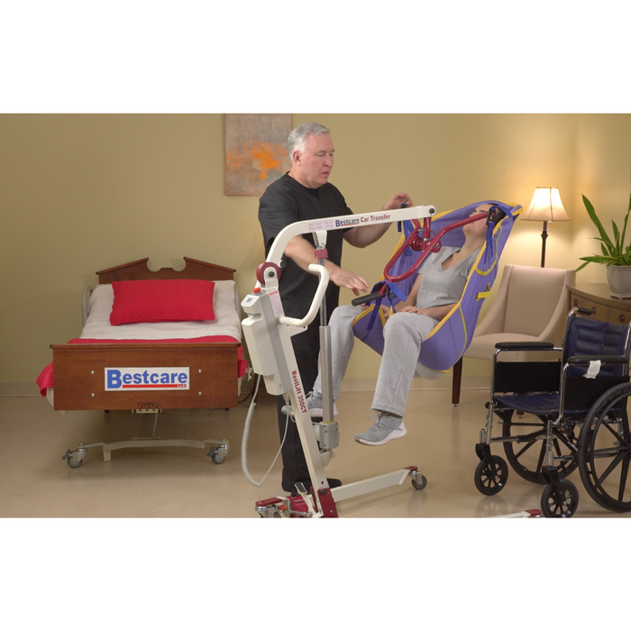 A man expertly operates a Bestcare BestLift PL350CT Folding Portable Electric Patient Lift, smoothly positioning an elderly person in a blue and yellow sling. Nearby, a Bestcare bed, wheelchair, and plant complete the scene, emphasizing the importance of portable mobility solutions.