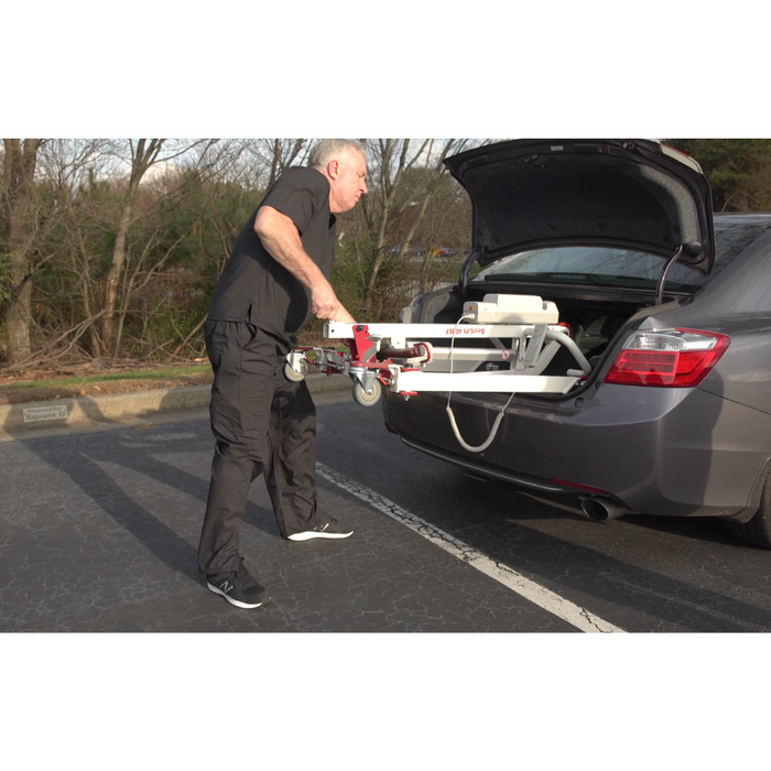 A person in black clothing places the Bestcare BestLift PL400EF Compact Foldable Electric Mobile Floor Lift into the open trunk of a gray car parked on a paved area. Trees and grass are visible in the background, showcasing this lifts portability for seamless adventures.