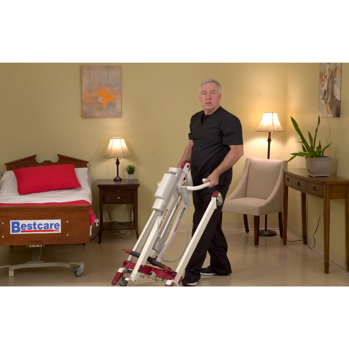 In a well-lit room featuring a bed, nightstand with lamp, and side table with plant, someone in black demonstrates the Bestcare BestLift PL400EF Compact Foldable Electric Mobile Floor Lift. A sign on the bed reads Bestcare.