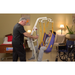 Using the Bestcare Bestlift PL400HE Electric Patient Lift, a caregiver skillfully transfers a seated patient in a purple sling. A wheelchair is nearby, with a bed in the background of the warmly lit home care setting.