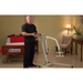 A man demonstrates a Bestcare Bestlift PL400HE Electric Patient Lift in a room with a hospital bed labeled Bestcare, ideal for home care. The setting includes a chair, table with lamp, and plant against beige décor, complemented by red and white bedding on the bed.