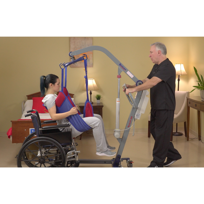 A man uses the Bestcare BestLift PL Series Full Body Electric Patient Lift to assist a woman from her wheelchair using a sling, safeguarding caregiver back health. In the care facility room, theres a bed, side table, and plant.