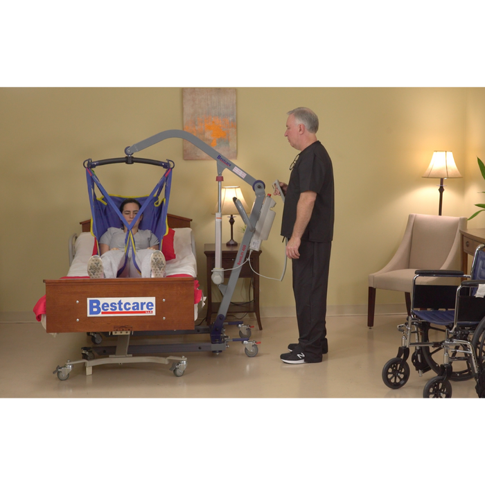 A caregiver uses a Bestcare BestLift PL Series Full Body Electric Patient Lift to assist someone in a blue sling into or out of bed, ensuring effective transfer and back injury prevention. The room has beige walls, a chair, wheelchair, and lamp.