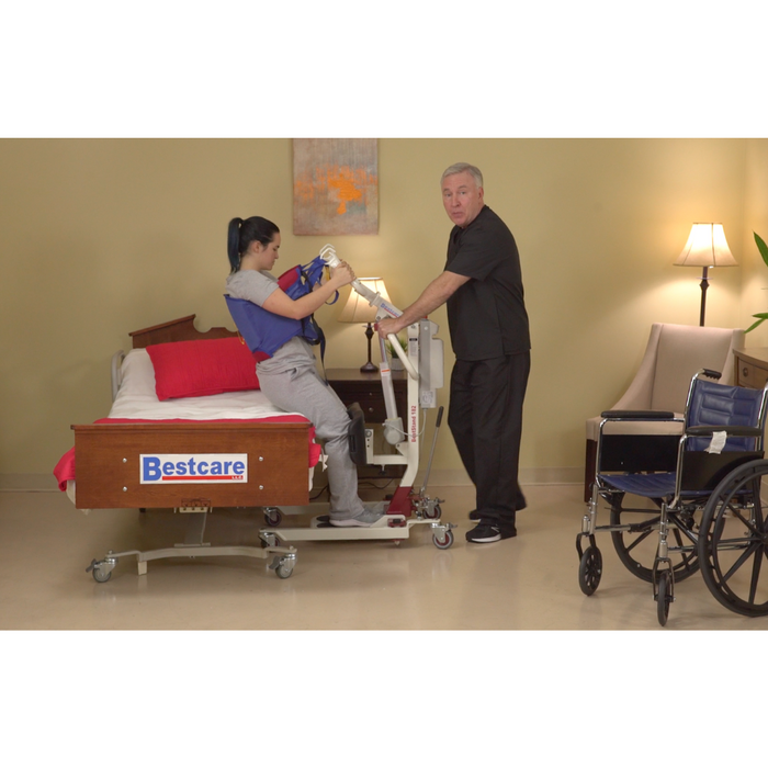 A caregiver uses a Bestcare SA Series Sit-To-Stand Compact Electric Patient Lift to assist a person on a hospital bed while transferring. The room includes a wheelchair, side table, and lamp, alongside the red and white Bestcare bedding that adds an organized touch to the clinical setting.