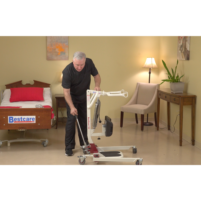 A man demonstrates the Bestcare SA Series Sit-To-Stand Compact Electric Patient Lift for seamless transfers beside a Bestcare bed in a room with a bedside lamp and plant on a table. The lift, with wheels, highlights its Medicare reimbursable benefits.