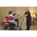 A man assists a woman using a Bestcare BestStand® SA Series Sit-to-Stand Patient Lift from her wheelchair. The room, with a bed, lamp, and wall art, offers a familiar environment. She is in a harness for the transfer. This setting resembles her home or care facility.