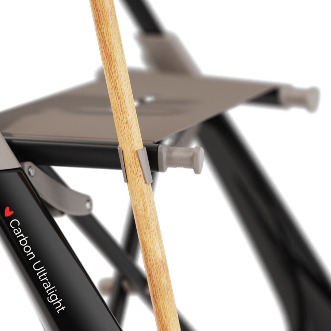 Close-up of a folding chair with "Carbon Ultralight" on the frame, showing a wooden walking cane clipped securely using the Cane Holder for ByAcre Ultralight Carbon Fiber Rollators, a handy accessory often paired with Carbon Rollators.