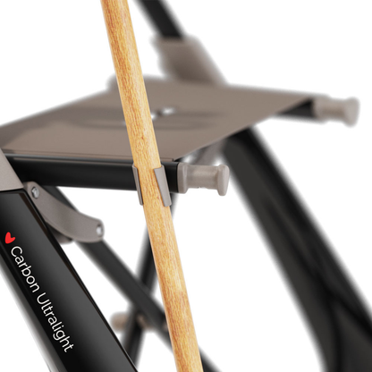 Close-up of a folding chair with "Carbon Ultralight" on the frame, showing a wooden walking cane clipped securely using the Cane Holder for ByAcre Ultralight Carbon Fiber Rollators, a handy accessory often paired with Carbon Rollators.