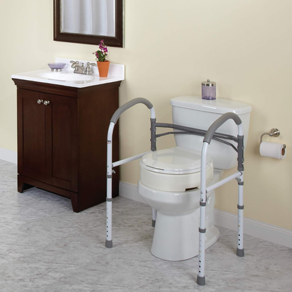 A bathroom features a white toilet with the Carex Bathroom Safety Rail, handicap grab bars, and a wooden sink vanity topped with a mirror, potted plant, soap dish, towel, and wall-mounted toilet paper.
