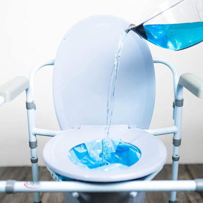 A close-up of a portable commode chair with a Carex Commode Liner with Absorbent Powder in the seat. A hand pours blue liquid into the liner, which sits on a wooden floor against a white background.