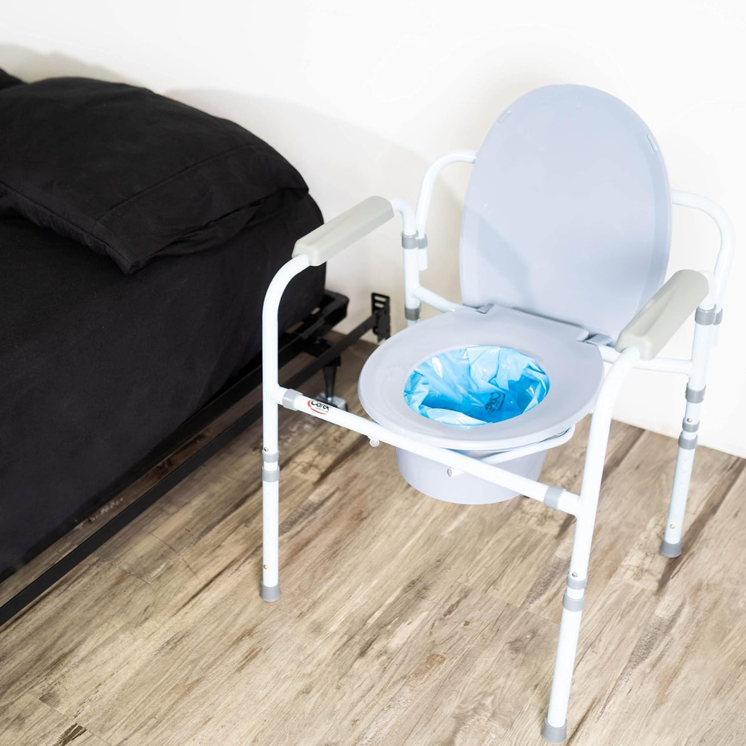 A portable bedside commode with a Carex Commode Liner featuring absorbent powder is next to a bed with black bedding on a wooden floor.