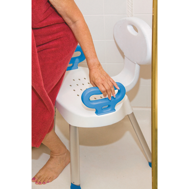 A person in a red towel stands in a tiled shower, holding the blue handle of the Carex E-Z Bath and Shower Seat with Handles, which is white with supportive handles and a backrest.