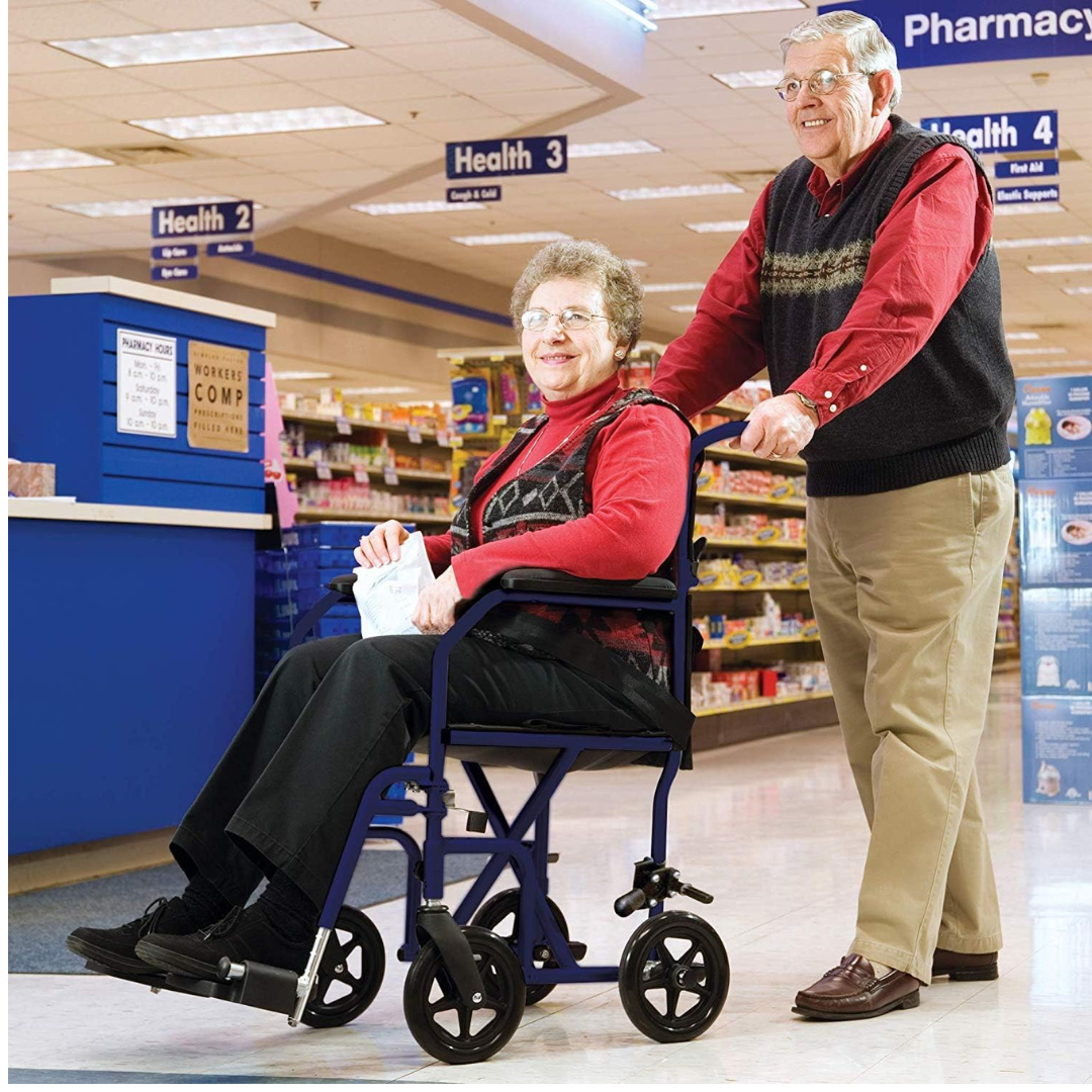 An elderly man smiles while pushing an elderly woman in a Carex Folding Steel Transport Wheelchair - 19" Seat through a pharmacy aisle. Both wear red tops and glasses, with health products on shelves in the background.