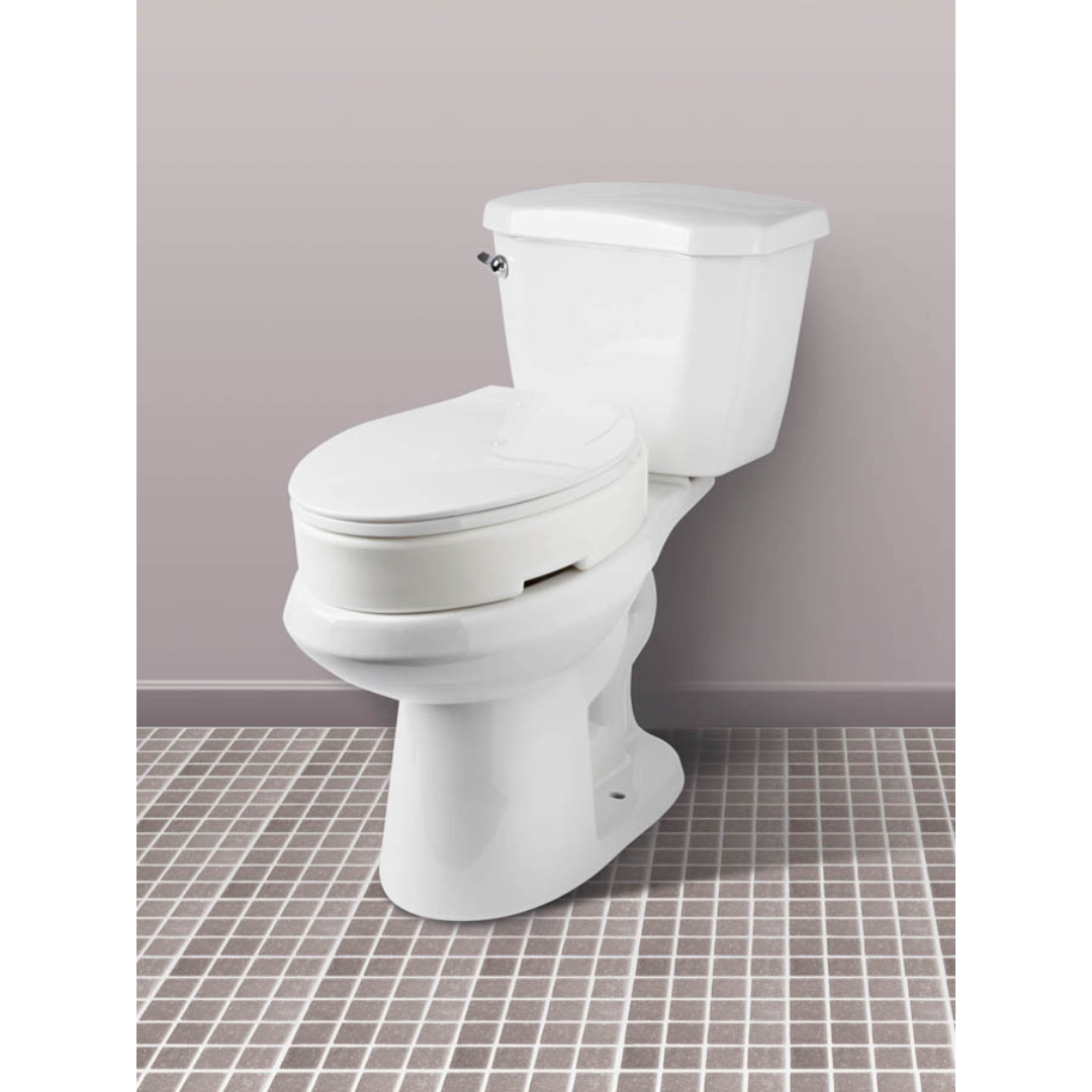 The Carex Hinged Toilet Seat Riser – Add 3.5 Inches sits on a gray and white tiled floor atop a white ceramic toilet with a closed lid, placed against a light purple wall.