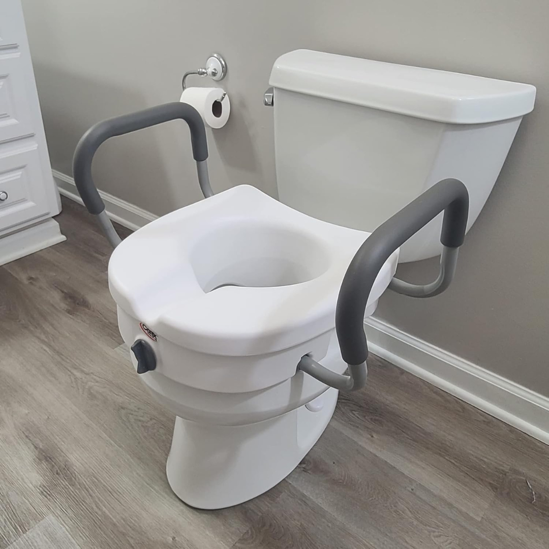 The Carex E-Z Lock 5" Raised Toilet Seat with adjustable gray armrests fits over a white toilet in a bathroom with light wood flooring and a toilet paper holder. The seat supports up to 300 lbs.