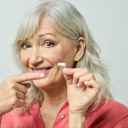 Smiling older woman with gray hair in a pink shirt holds and points to the Ceretone Core One Pro Hearing Aids - Invisible-In-Canal, set against a plain light background.