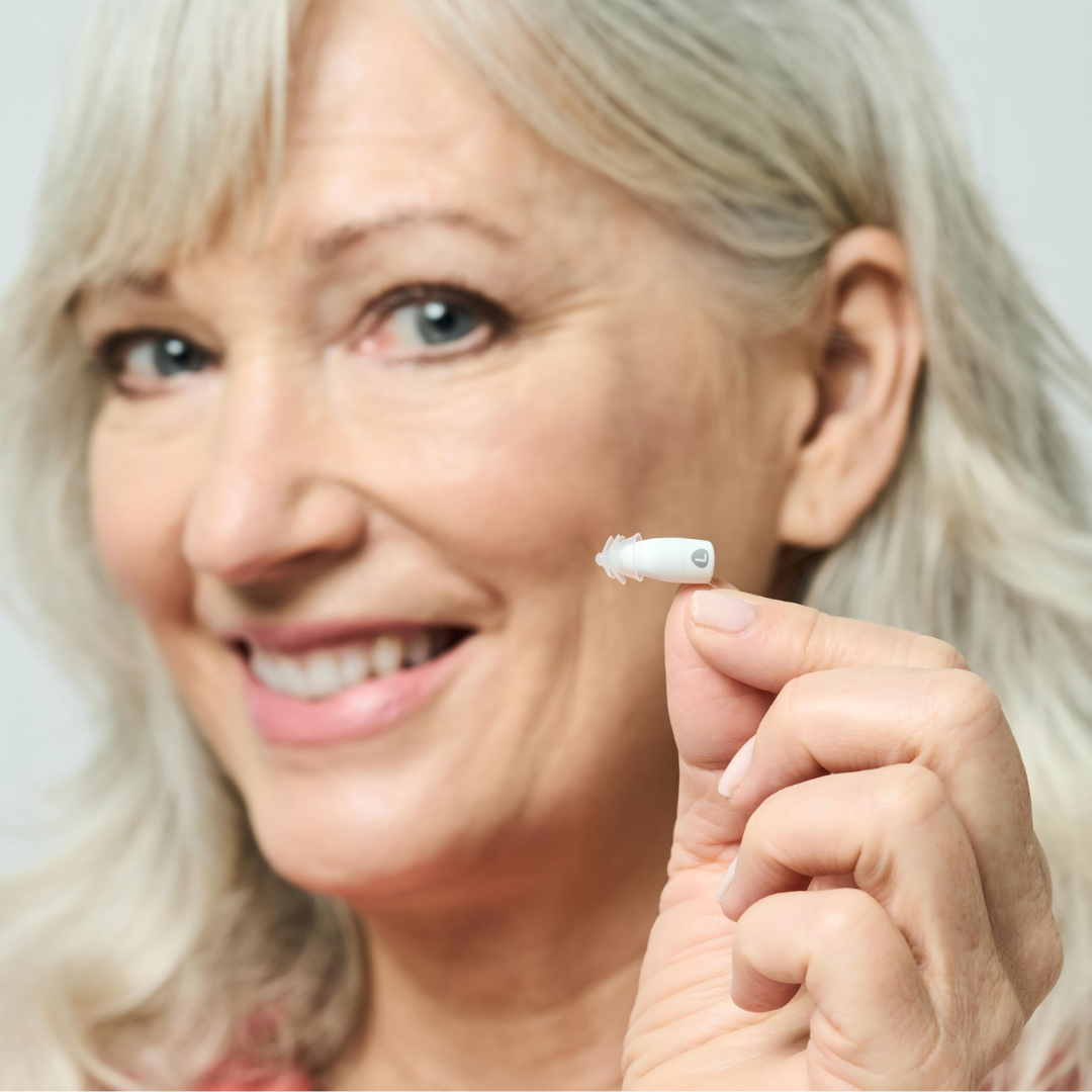 A smiling older woman with gray hair holds a Ceretone Core One Pro Hearing Aid (Invisible-In-Canal) between her fingers, presenting it to the camera against a softly blurred, neutral background.
